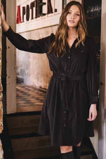 Kvinde med gyldent langt hår, læner sig op af en væg, iført en knælang brun kjole med bælte i taljen